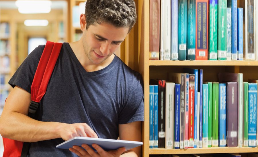 Student leaning against bookshelf holding a tablet pc at the library